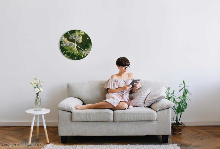 Eine Frau sitzt auf einer Couch mit einem Buch in der Hand und einer Uhr im Hintergrund hinter sich, moosbild, mooswand, moos pflanzen, moos, moos deko, moos art